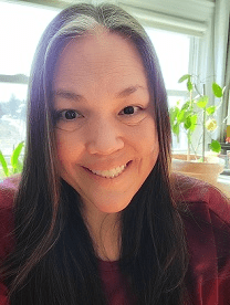 Woman smiling warmly at the camera, surrounded by sunlight and potted plants, suggesting a cozy indoor setting. Relevant for personal or lifestyle content.