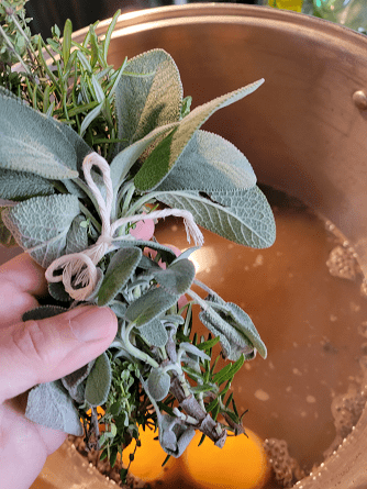 A hand holds a bundle of fresh herbs—sage, thyme, and rosemary—above a pot of simmering liquid with oranges, preparing for cooking.